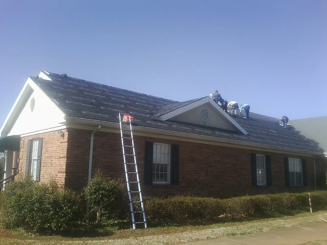 Crew tearing shingles off a brick ranch roof with a ladder set — Emergency Roof Repair work by our crew in Bellerose