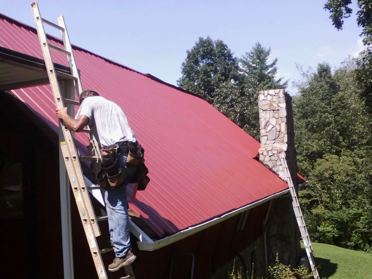 Roofer on a ladder beside a red metal roof and stone chimney — Soffit & Fascia Repair by Roofing Brothers in Bellerose